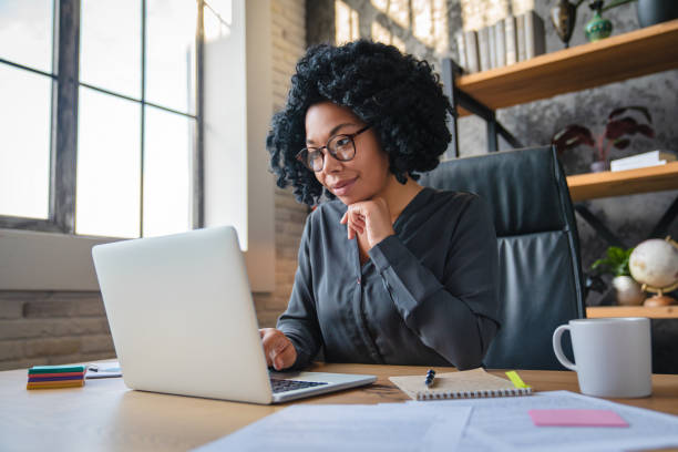 Happy young African American woman businesswoman manager in casual formal wear eyeglasses looking at computer screen reading email working distantly on online project at modern home office. stock photo