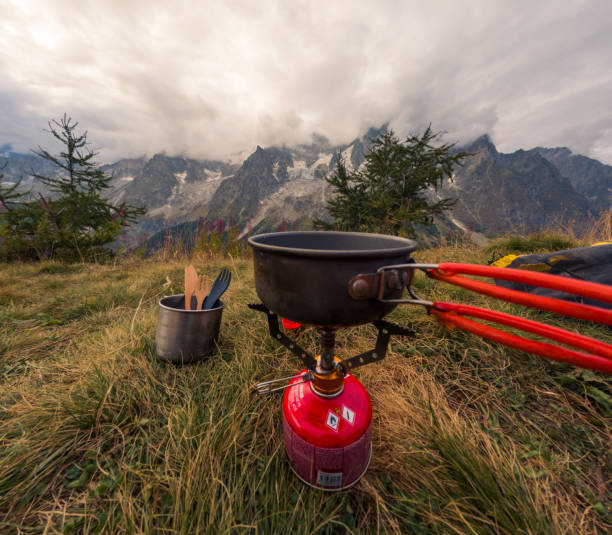 A camping stove with a pot on a grassy alpine campsite, set against a breathtaking mountain range under a cloudy sky.