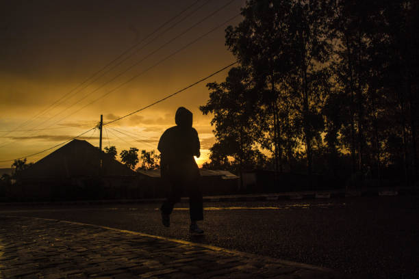A dramatic silhouette of a person walking along a cobblestone path, framed by the soft glow of a golden sunrise. The interplay of light and shadow highlights the serenity of an early morning, with trees and rooftops faintly visible in the background. This captivating shot perfectly captures the mood of peaceful solitude and the promise of a new day, ideal for storytelling or contemplative visual narratives.
