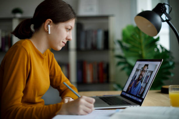 Teenage girl with wireless headphones taking notes while watching an online tutorial on her laptop at home