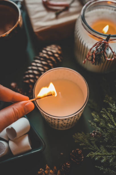 istock A hand lighting a festive candle with a matchstick, surrounded by pinecones, marshmallows, and evergreen branches, creating a warm and cozy Christmas atmosphere 2185949949