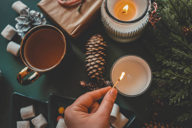 istock A hand lighting a festive candle with a matchstick, surrounded by pinecones, marshmallows, and evergreen branches, creating a warm and cozy Christmas atmosphere 2185949952