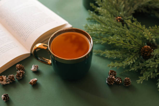 A cup of tea on a green surface with an open book, pinecones, and evergreen branches, creating a cozy and festive holiday atmosphere. Warm light and seasonal decor enhance the mood