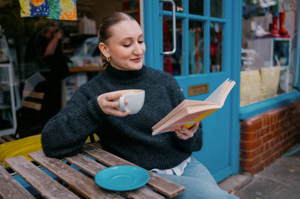A young woman sits outside of a bright and cosy coffee shop reading a book while enjoying her coffee