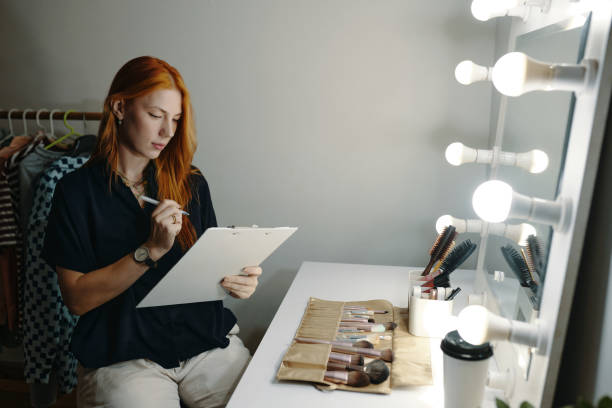 istock Sitting Female Makeup Artist Holding Clipboard by Vanity Mirror 2185952427
