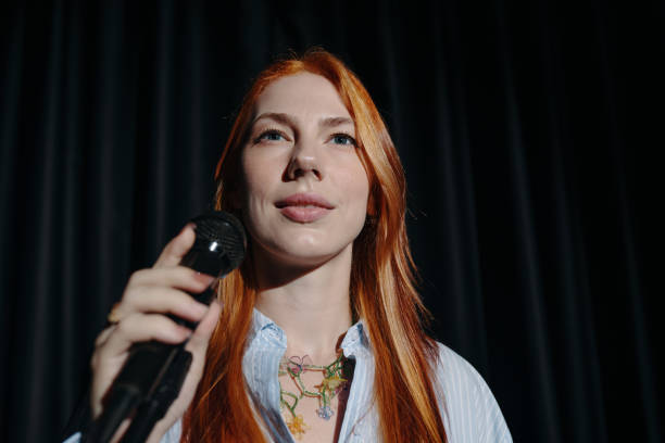 Portrait of woman with long red hair holding microphone while standing on stage against dark backdrop, showing engaged expression and casual attire