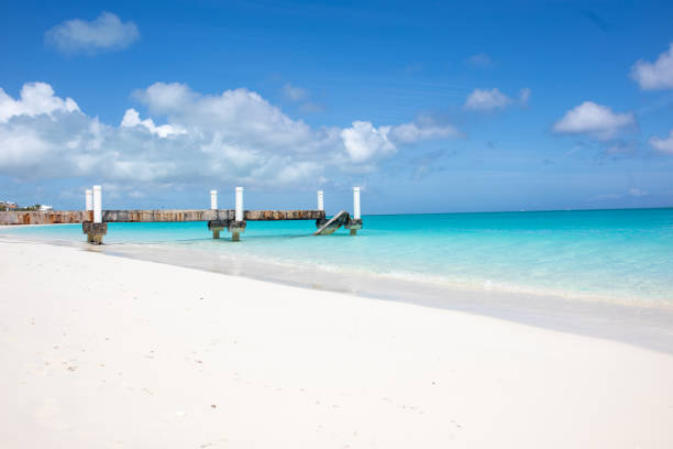Partially collapsed concrete pier extending into shallow crystal clear turquoise water . The pier has white cylindrical pillars a that contrast against its weathered and rusty concrete frame. The foreground shows pristine white sand, while the vibrant blue sky with clouds complements the tropical setting.