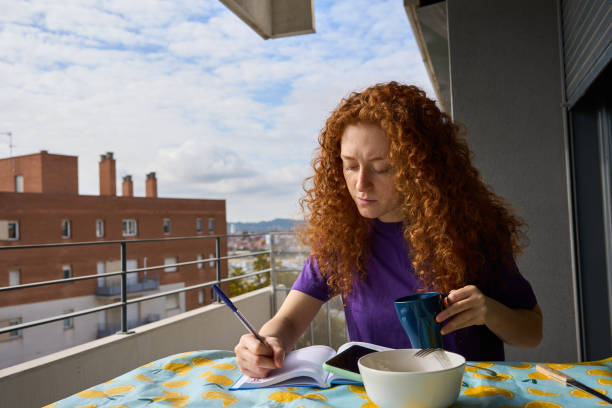 Young woman with red, curly hair enjoys breakfast and writes on her balcony in barcelona, reflecting in a cozy city setting