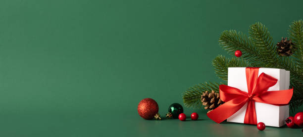 istock A beautifully wrapped Christmas gift with a red ribbon surrounded by pine branches and baubles, set against a green background, evoking holiday spirit and joy 2185957253