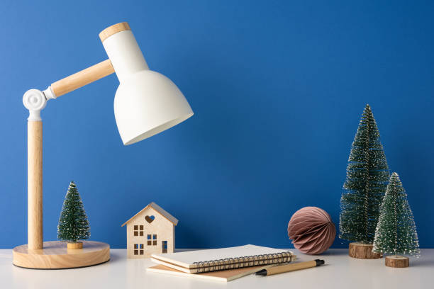 A side view minimalist winter-themed desk with a Scandinavian design, featuring a lamp, notebook, wooden house, and mini Christmas trees against a blue background