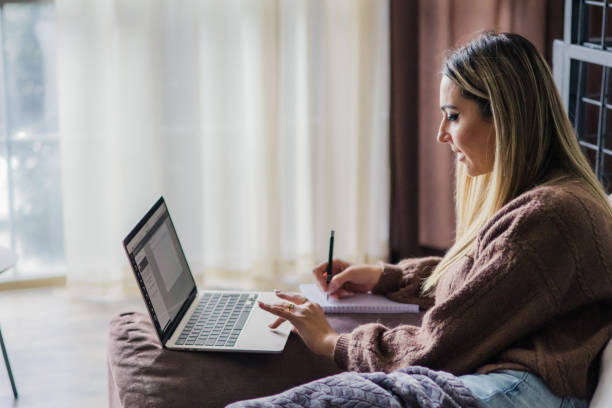 istock Beautiful young woman is working on laptop at home. Making notes in notebook while investing and buying at crypto market. Calculating profit from sales. 2185962078