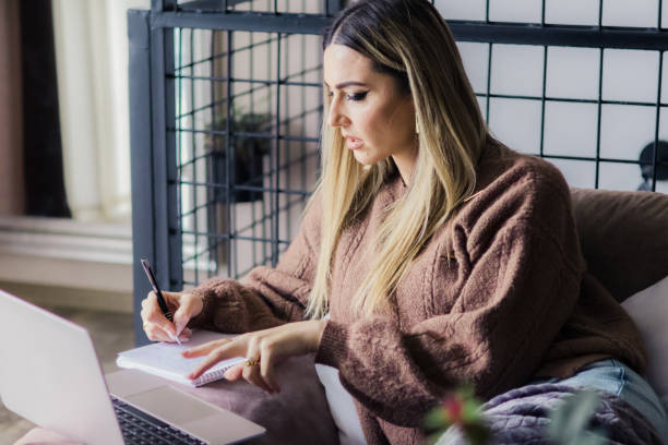 istock Beautiful young woman is working on laptop at home. Making notes in notebook while investing and buying at crypto market. Calculating profit from sales. 2185962311