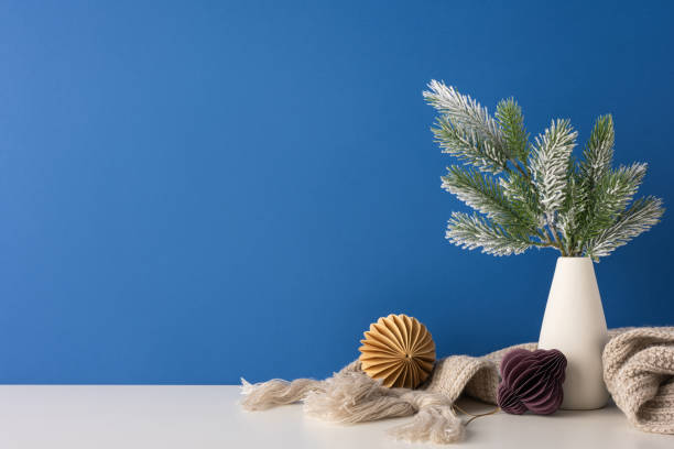 istock Minimalistic Scandinavian-style side view decor featuring a frosted vase with pine branches, accompanied by paper ornaments and a cozy scarf on a white surface 2185975693