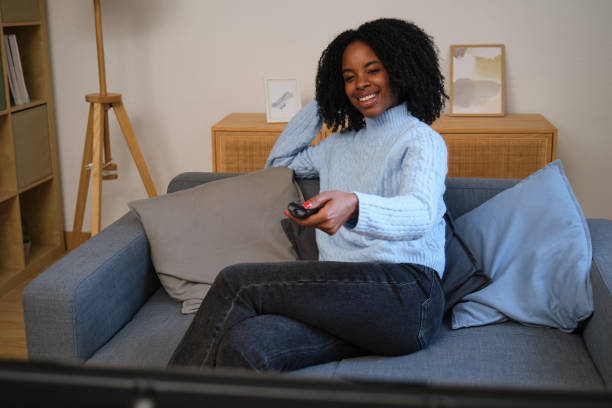 istock Young woman sitting on sofa watching television and holding remote control 2185982974