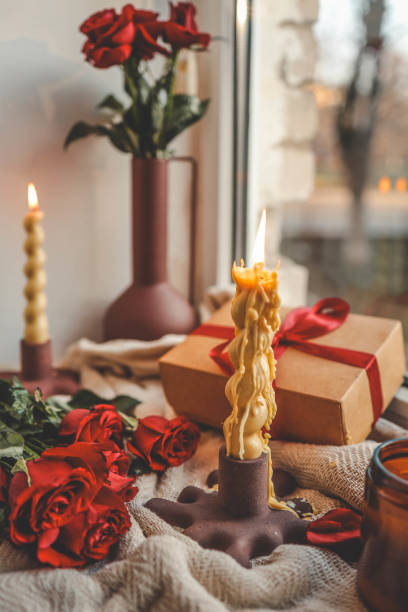 istock A cozy window-side scene featuring a lit beeswax candle with a unique shape, vibrant red roses, a gift box with a ribbon, and a soft textured fabric, creating a warm and serene ambiance 2185989186