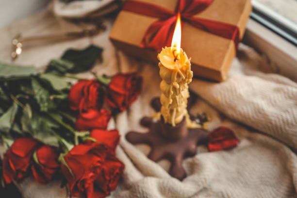 istock A cozy window-side scene featuring a lit beeswax candle with a unique shape, vibrant red roses, a gift box with a ribbon, and a soft textured fabric, creating a warm and serene ambiance 2185989188