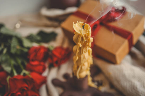 A recently extinguished beeswax candle with smoke curling upwards, surrounded by vibrant red roses and a gift box tied with a ribbon in a cozy and warm setting.