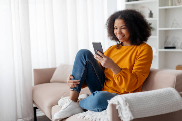 istock Relaxed young African American woman smiling and browsing her smartphone while reclining on couch, happy millennial lady using mobile phone for online communication or shopping, resting in living room 2186008694