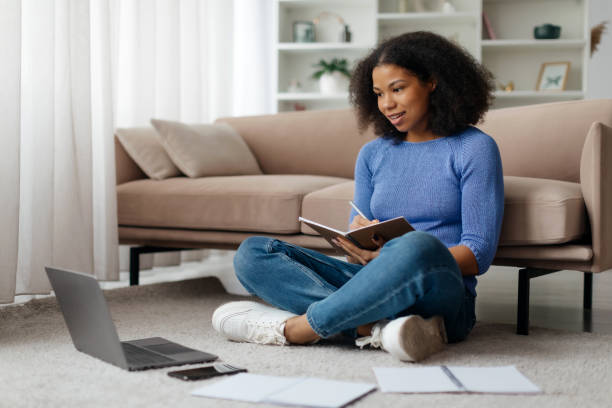 istock Smiling young African American woman working on laptop with documents aside, happy eastern freelancer lady sitting cross-legged on floor in living room, fully immersed in her tasks at home, copy space 2186011250