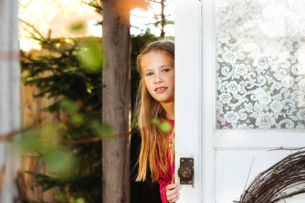 A beautiful, eight-year-old girl smiles at the camera as she peeks out from behind a door on a  rustic porch. Shot with a Canon 5D Mark IV.