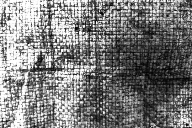 Black and white close-up of a textured woven fabric or material, showcasing a grid-like pattern with visible imperfections, folds, and shadows. The interplay of light and dark adds depth to the composition