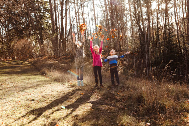 Twin girls toss leaves in the air with their little brother outside on a beautiful, autumn day.