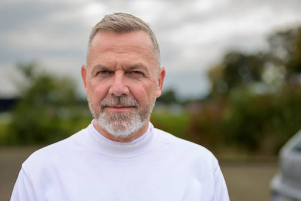 A close-up portrait of a mature man with a gray beard and short hair, wearing a white shirt.