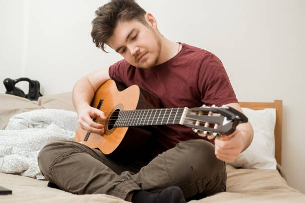 istock A young man is sitting on the bed and tuning his guitar. 2186071421