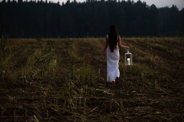 A woman in a white dress holding a lantern walks through a misty forest at dusk, creating a mystical atmosphere.