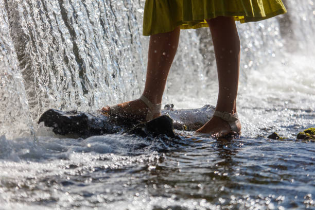 A woman is standing on a rock in a river. The water is flowing and splashing around her feet. The scene is peaceful and serene, with the sound of the water