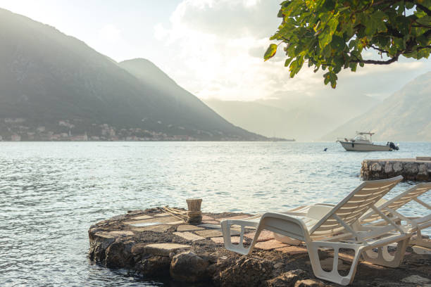 istock A tranquil seaside spot with lounge chairs on a stone pier, overlooking calm waters, majestic mountains, and a small boat in the distance. 2186097877
