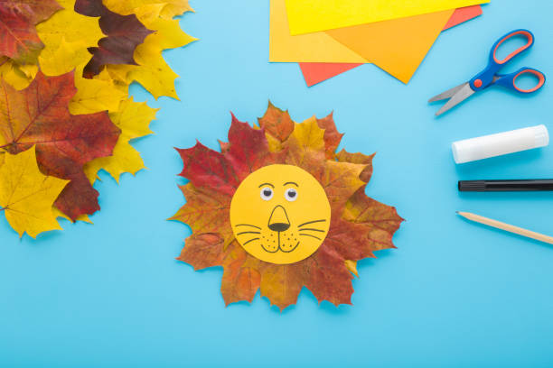 istock Beautiful smiling lion head shape created from colorful maple leaves and paper on light blue table background. Pastel color. Making autumn decorations. Closeup. Top down view. 2186129455