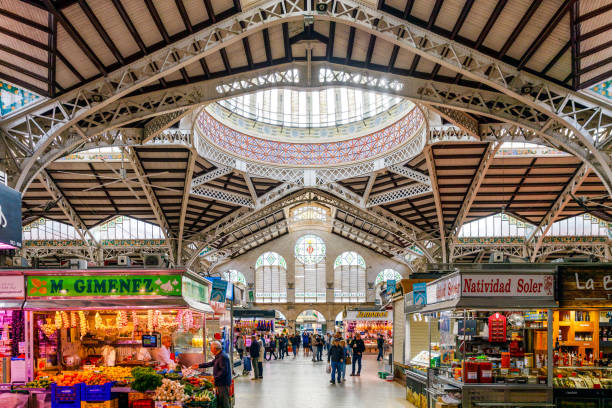 Valencia, Spain. Monday,  18th of November 2024. The photo captures a vibrant scene inside the iconic Central Market of Valencia, Spain. Known locally as Mercado Central, this historic market is one of Europe’s oldest functioning food markets and a landmark of modernist architecture. The interior features a stunning combination of historical charm and contemporary functionality, with colorful stalls offering fresh produce, meat, fish, and regional delicacies. Natural light streams through the market's glass windows, highlighting the intricate ironwork and decorative ceramic tiles. This image perfectly encapsulates the dynamic atmosphere of this cultural hub, making it ideal for travel publications, culinary blogs, or architectural features.