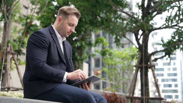 istock Businessman sitting at park while working on tablet by reading report. Urbane 2186132125