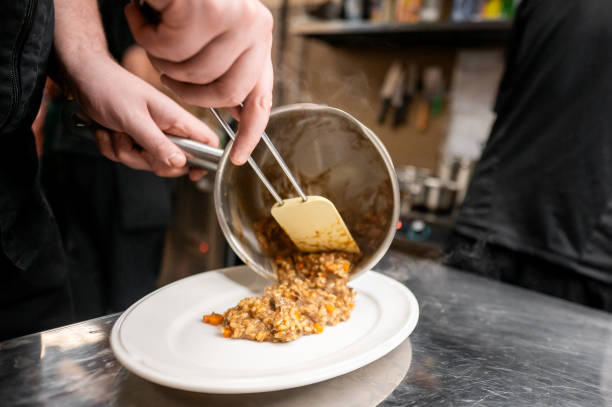 A close-up of hands pouring a risotto from a bowl onto a white plate, likely preparing or plating a meal. The scene suggests a culinary process, emphasizing the art of cooking and food presentation.