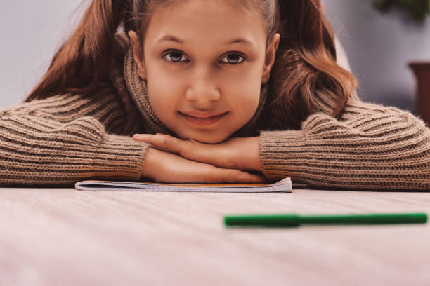 Portrait of a smiling elementary age girl resting her head on her hands on top of her notebook after finishing her homework, with a green pen in the foreground