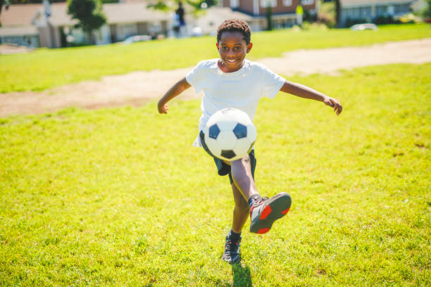 Cute elementary age boy dressed in a sports uniform (blue t-shirt and black shorts). He is posing on a green artificial turf carpet with the soccer ball. Studio shooting on a black background