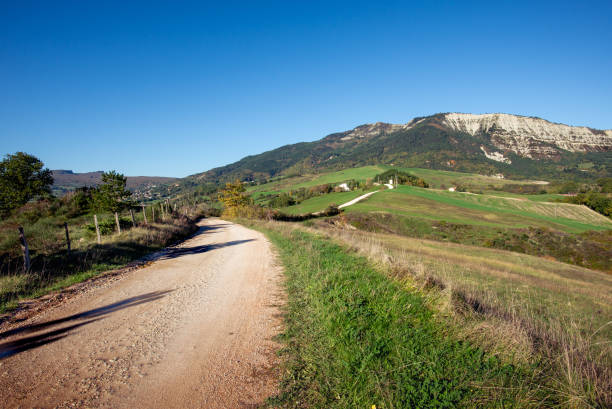 Serene landscape of rolling grassy hills accented with rocky outcrops. In the background, the vast expanse of mountains and valleys unfolds under a clear blue sky, creating a breathtaking panorama. The golden hues of the sunlight, captured during sunset, enhance the warmth and beauty of the terrain. This photo was taken on Grand Colombier famous summit in Bugey mountains, in Ain, Auvergne-Rhone-Alpes region in France with a bright beautiful sunset in august.