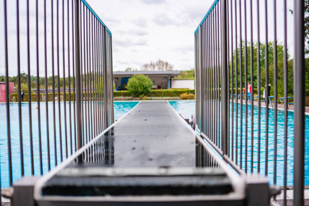 A wet diving board leading into a large outdoor swimming pool with protective railings, surrounded by greenery and overcast skies, suggesting a recreational day at a public pool.