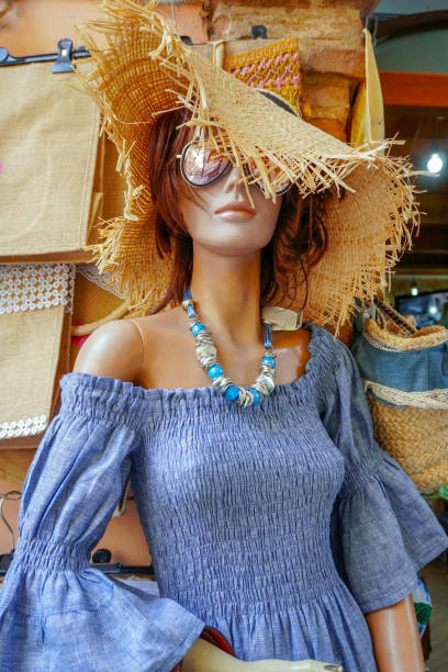 A photo of a mannequin dressed in a boho style. The mannequin is wearing a wide-brimmed straw hat with frayed brim, large sunglasses and a blue off-the-shoulder dress with voluminous sleeves.