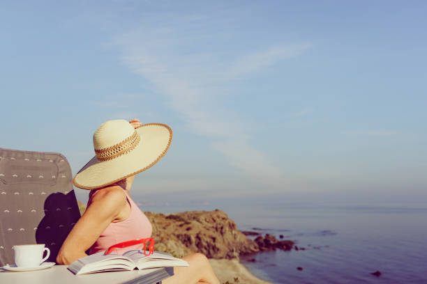 Mature tourist woman relaxing on coast with book and coffee cup, enjoy sea view from cliff. Reading on holidays. Relax time.