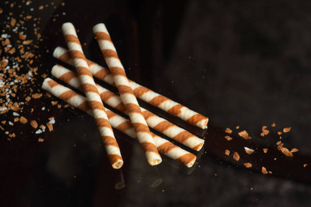 Five striped wafer sticks lying diagonally on a reflective surface, surrounded by scattered crumbs, showcasing a playful scene of delicacy and snack time enjoyment.