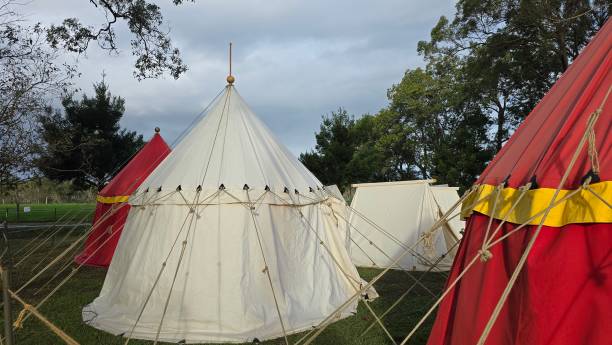 istock Medieval-style camp with white and red canvas tents, secured with ropes and poles, set on a grassy field surrounded by trees under a cloudy sky. 2186326624