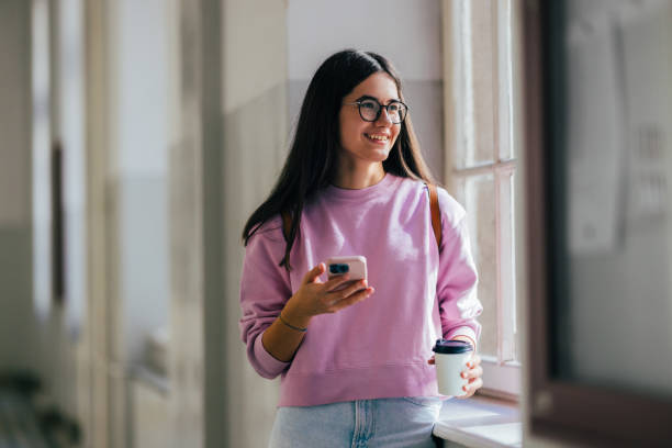 Smiling Young Woman Holding Smartphone and Coffee Cup by Window