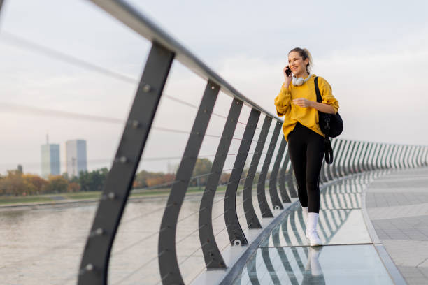 istock Cheerful woman strolls along a modern bridge, chatting on her phone while enjoying the serene cityscape at dusk by the water 2186389621
