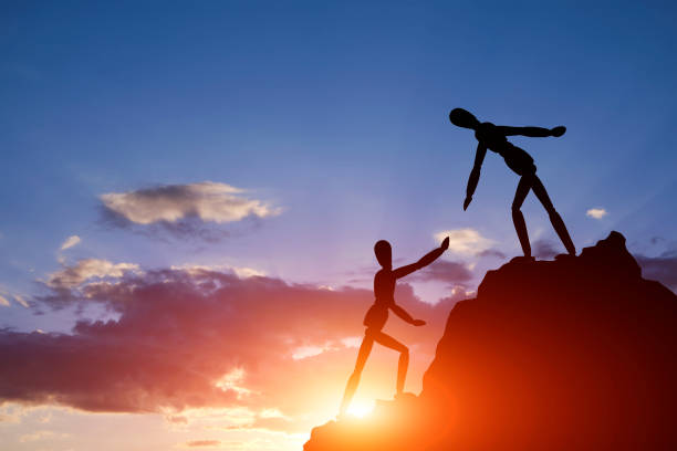 istock Two silhouettes mannequin climb mountain, one helping other, extending helping hand. Against background sky, sunset and clouds. Concept of help and success in teamwork. 2186402931