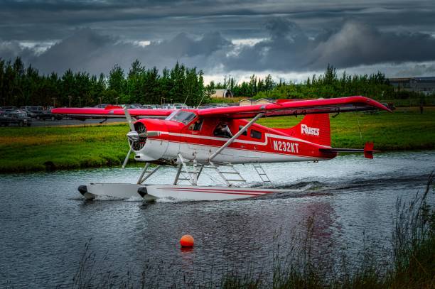 Anchorage, United States – July 24, 2024: A red and white seaplane on a body of water with a cloudy sky and green trees in the background.