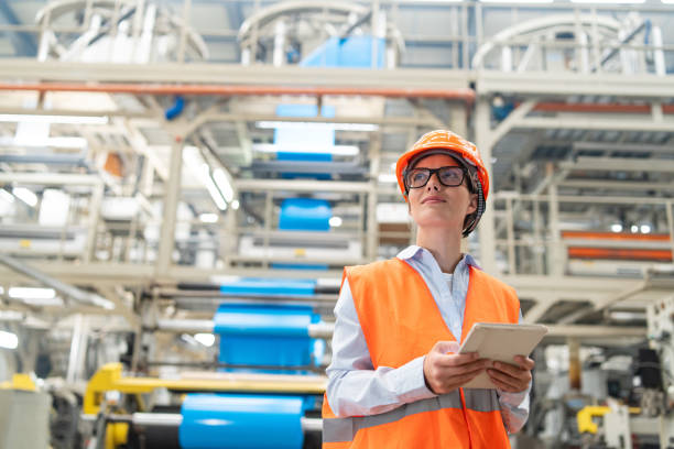 Engineer overseeing operations on tablet in bright industrial plant wearing safety gear.