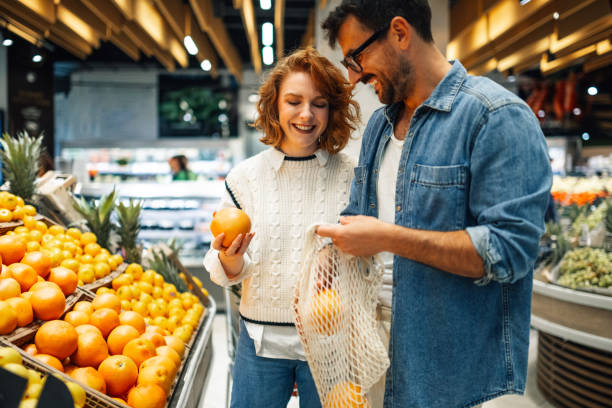 счастливая пара выбирает апельсины в многоразовой сетчатой сумке в продуктовом магазине - товар стоковые фото и изображения