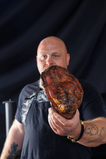 istock A rugged, bald, bearded butcher holds a large pork knuckle in his hands, set against a dark background. themes of traditional meat preparation, craftsmanship, and culinary expertise. 2186433409